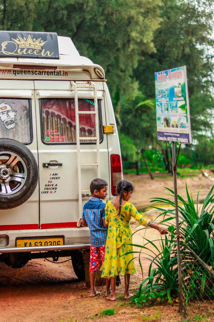 Two children stand near a camper van on a tropical trail, exploring their surroundings.