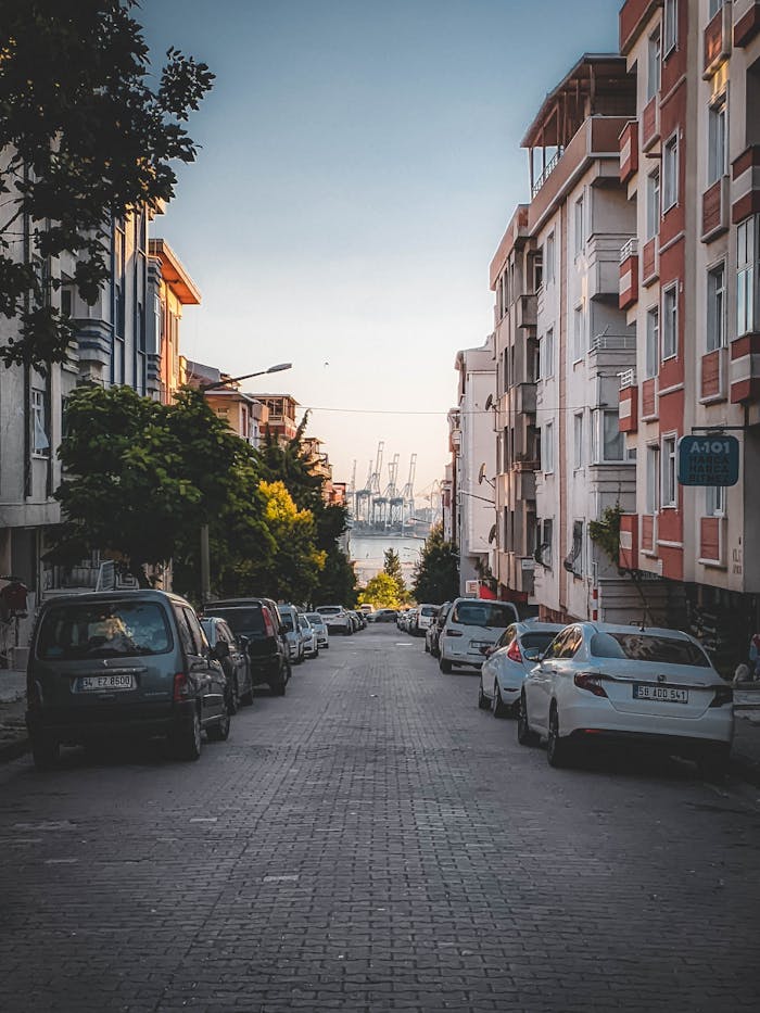 Picturesque urban street with cars parked alongside, leading to a distant harbor with cranes and a serene sky.